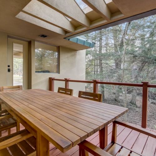 A covered patio with a wooden dining table and chairs, looking out over a wooded forest with tall trees.