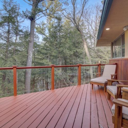 A wooden deck with red railing, patio chairs, and a view of a forested backyard under clear blue sky.