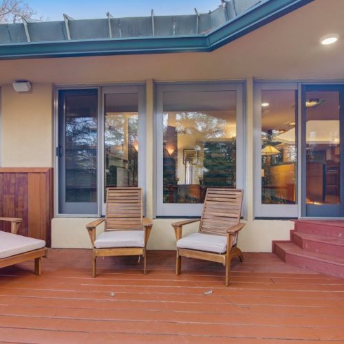 A patio lounge area with two wooden chairs, tiled floor, and glass doors under an overhang.