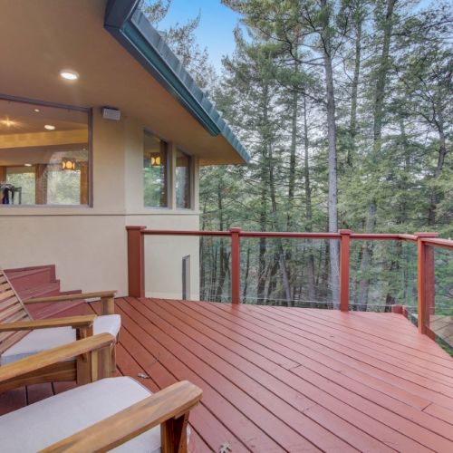 A cozy wooden deck with red railing and lounge chairs, attached to a house, overlooking tall pine trees in a forest.