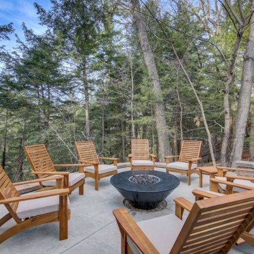 A wooden patio set with lounge chairs surrounds a round fire pit, nestled in a wooded outdoor setting.