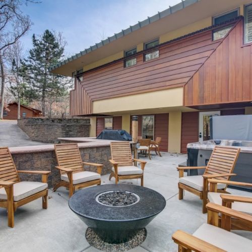 A modern apartment patio with wooden lounge chairs, a round fire pit, and a covered hot tub area in front of the building.