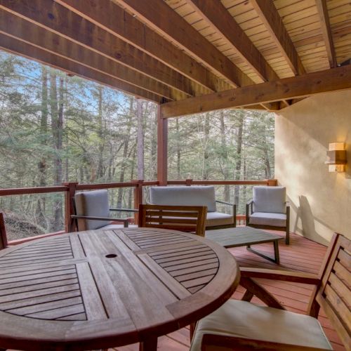 A covered wooden deck patio with a round table, cushioned chairs, and a view of the forest through tall windows.