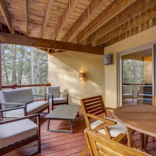 A covered wooden deck with outdoor seating&mdash;cushioned chairs and tables&mdash;overlooking a forested yard, with a glass door in view.