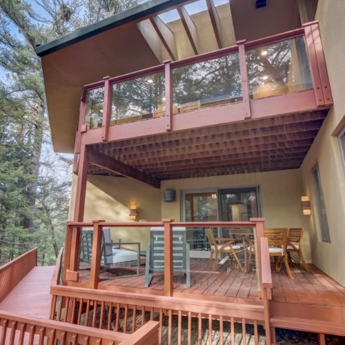 A modern house balcony with pink railings, glass panels, outdoor furniture, and a forest view behind it, outdoors.