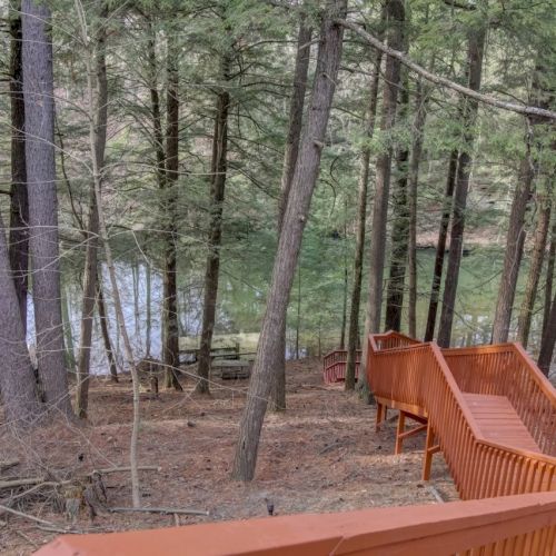 A wooded campsite with an orange deck and stairs beside a calm lake visible through the trees.