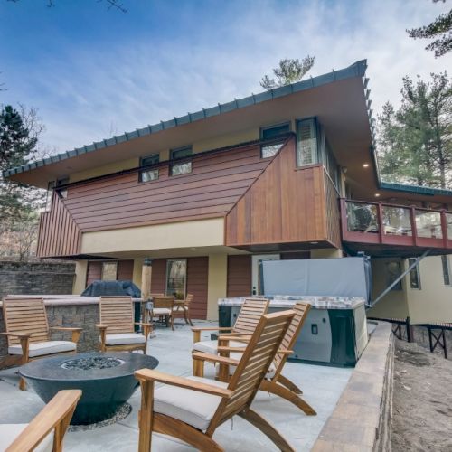 A modern house with a spacious patio, hot tub, wooden chairs, and a red deck railing surrounded by trees and sky.