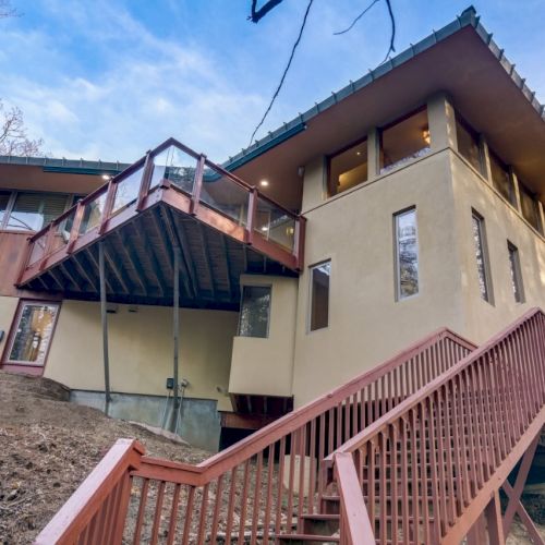 A modern multi-level house exterior with a red wood deck railing, glass balcony, and large windows under a blue sky.