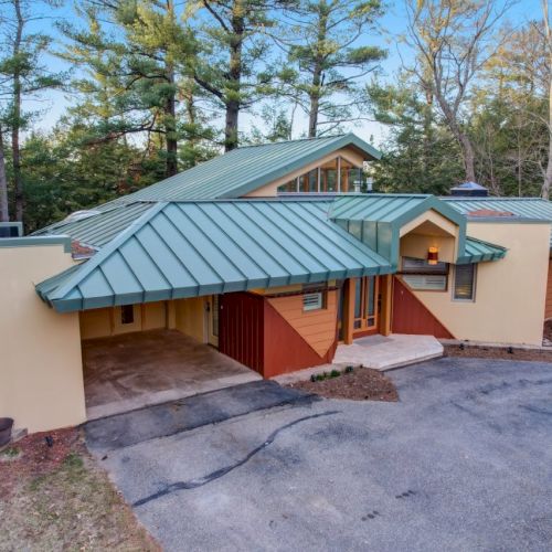 A modern house with a green metal roof, red accents, and a driveway surrounded by trees in a wooded setting.