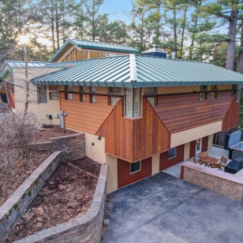 A modern cabin with a blue metal roof, wood siding, and a stone patio nestled among tall pine trees.