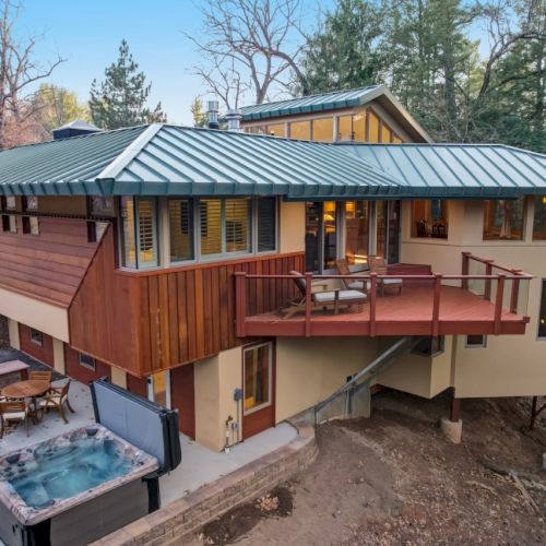 A modern cabin with a green metal roof, a deck with chairs, and a hot tub in the courtyard, surrounded by trees.