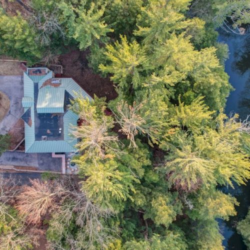 Aerial view of a rural house beside a winding road, surrounded by dense green trees and a small river.
