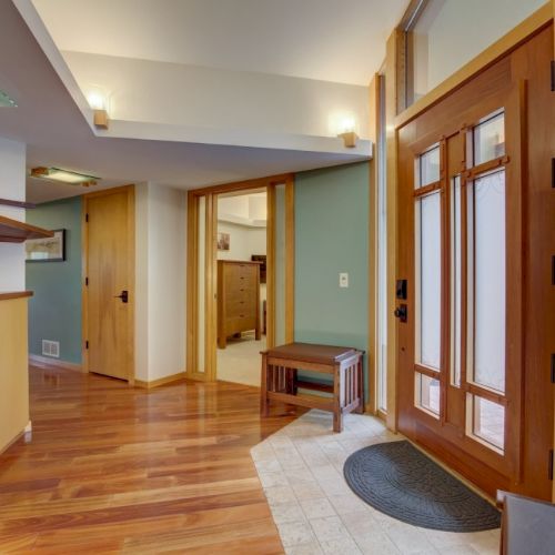 A bright interior entryway/hall with polished wood floors, wooden doors and windows, a bench, and a dark round floor mat.