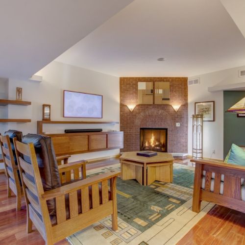 A cozy living room with a brick fireplace, wooden furniture, patterned rug, and a large windowed arch near a seating area.