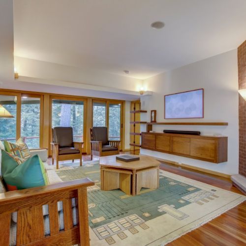A cozy living room with wooden furniture, large windows, a fireplace, and a patterned rug.