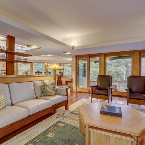 A cozy lodge-style living room with a sofa, two leather chairs, a wooden coffee table, and large forest-view windows.