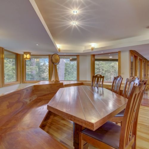 A bright wooden dining hall with a long table, cushioned chairs, large windows, and ceiling spotlights.