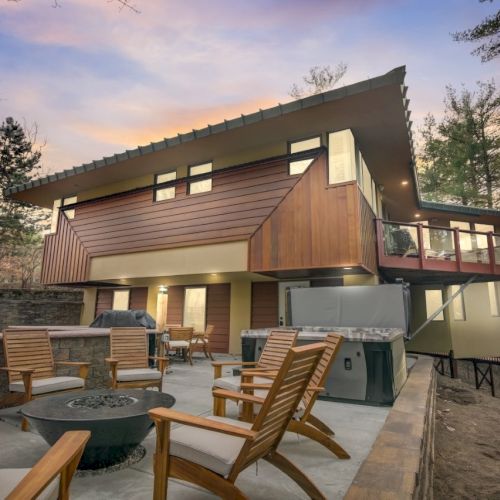 A modern house patio at dusk with wooden deck, outdoor chairs, a fire pit, and a hot tub in the yard.
