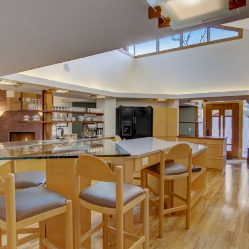 A bright modern kitchen with a central island, bar stools, stainless appliances, and glassware shelves under warm ceiling lights.