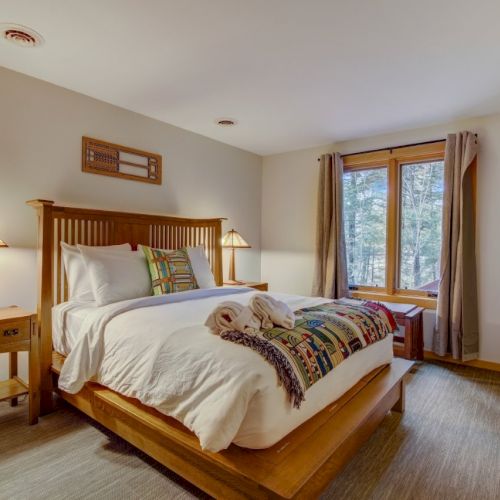 A cozy bedroom with a wooden bed, lamps, patterned pillows, and a window with curtains overlooking trees.