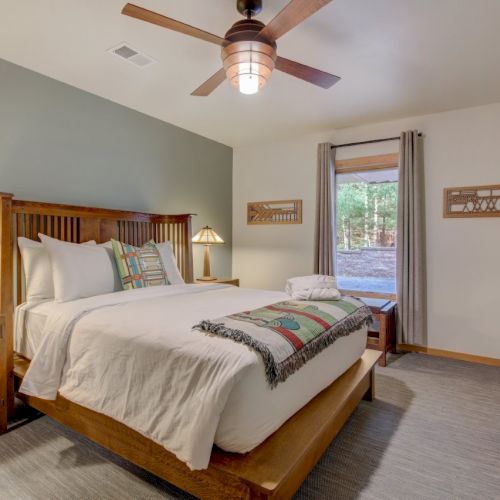 A bright bedroom with a wooden bed, ceiling fan, bedside lamps, and a window with curtains and a view outside.