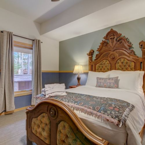 A bright bedroom with an ornate wooden bed, bedside lamps, patterned quilt, and tall curtains by a window.
