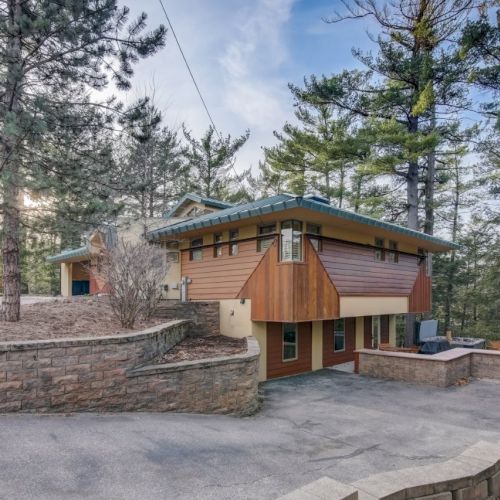 A modern house with a metal roof in a wooded area, with stone walls and a paved driveway winding through trees.