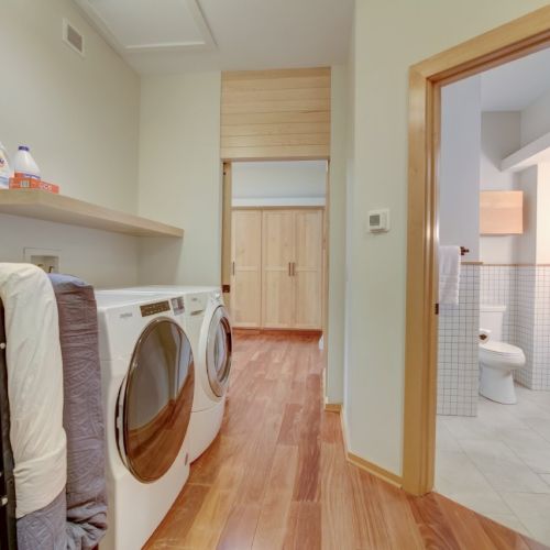 Bright laundry room: washer/dryer, detergent on a shelf, and a doorway into a tiled bathroom with a toilet.