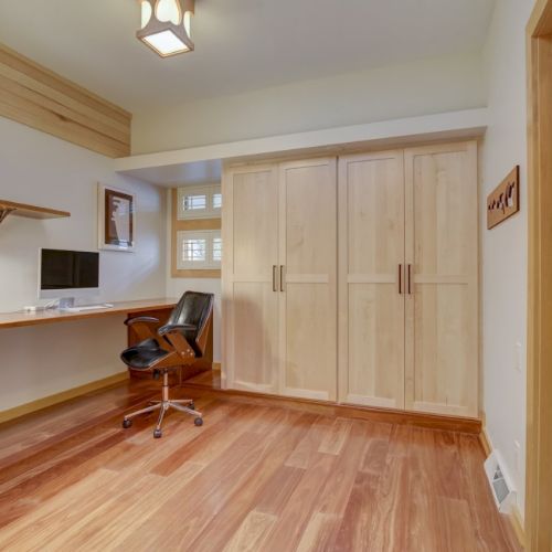 A bright office with wooden built-in cabinets, a swivel chair, pendant lights, and a doorway to a tiled room.
