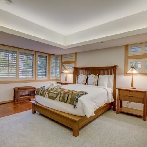 A bright bedroom with a wooden bed, pillows, bedside tables, lamps, and large shuttered windows.