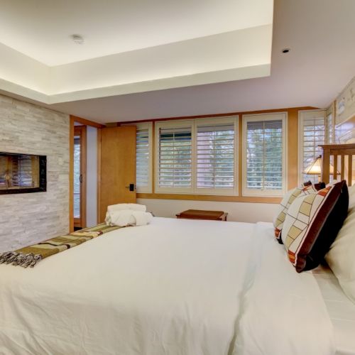 A bright modern bedroom with a white bed, stone fireplace wall, and large windows with blinds.