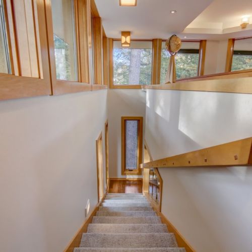 An indoor staircase with gray carpet, wooden railings, and large windows overlooking trees, in a bright, modern home.