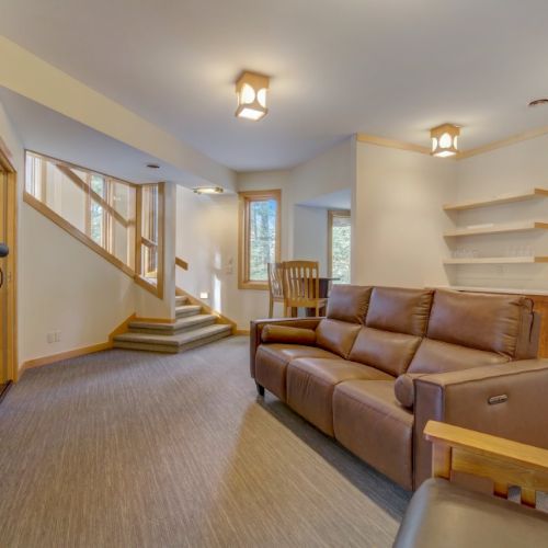 A modern living room with a brown leather sofa, wood accents, a stone fireplace, and an open staircase under warm lights.