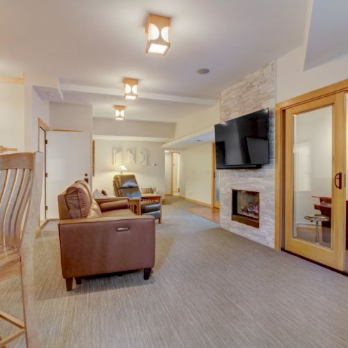 Bright living room with leather seating, a wall TV over a fireplace, and a dining counter under warm ceiling lights.