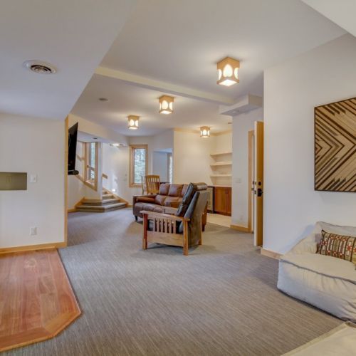A bright living room with gray carpet, leather chairs, a small sofa, warm ceiling lights, and large geometric wall art.