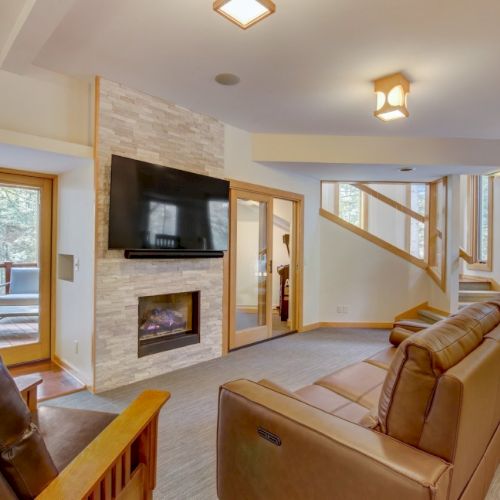 A bright living room with a stone fireplace, wall-mounted TV, leather recliner sofas, and large windows.