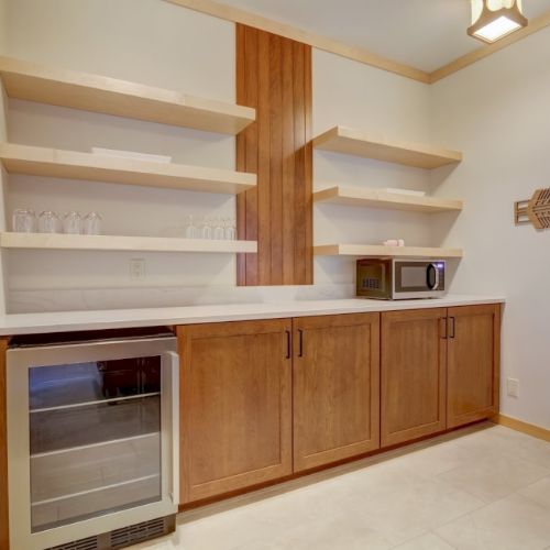 A modern kitchenette with wooden cabinets, open shelves, glassware, a fridge, and a microwave in a tidy room.