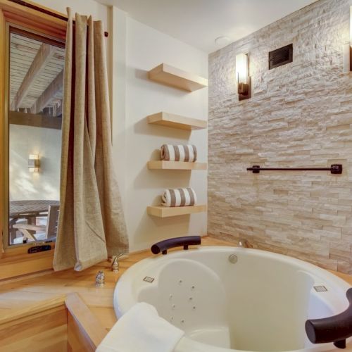 A modern bathroom with a stone-tile wall, towel shelves, and a large bathtub beside a window with curtains.
