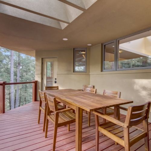 A covered balcony dining area with a long wooden table and chairs, overlooking tall forest trees through windows, in daylight.