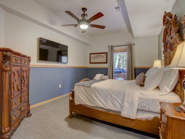 Bright bedroom with an ornate wooden bed, wall TV, ceiling fan, dresser, and lamps.