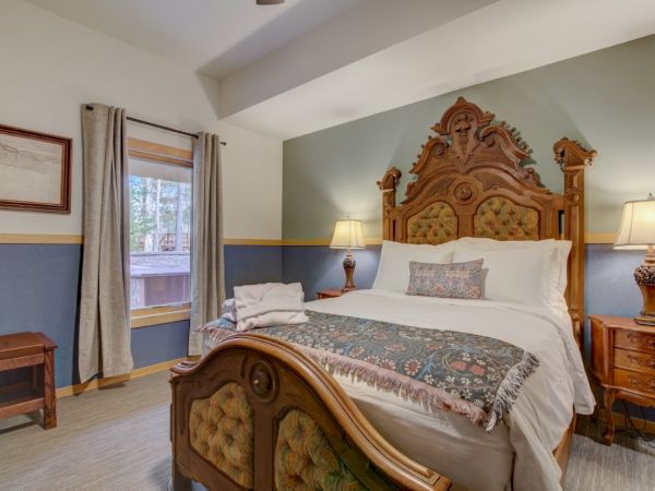 A warmly lit bedroom with an ornate wooden bed, bedside lamps, a dresser, and curtained window, neatly made.