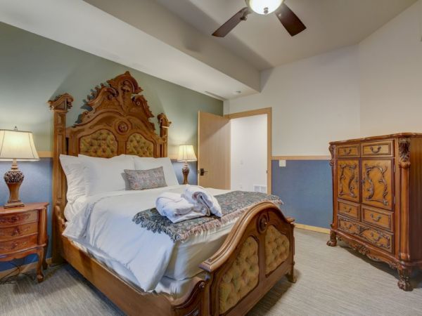 A stylish hotel bedroom with an ornate wooden bed, bedside lamps, ceiling fan, and a decorative dresser with mirror.