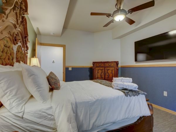 A hotel-style bedroom with a large bed, ornate headboard, dresser, ceiling fan, and wall-mounted TV.