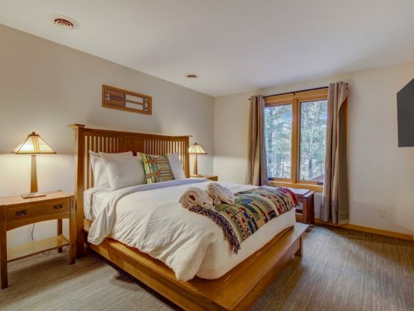 A cozy bedroom with a wooden bed, bedside lamps, pillows, curtains over a forest-view window, and a wall TV.