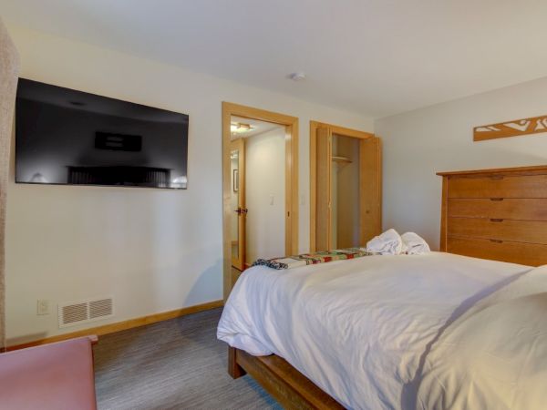 A bright bedroom with a wooden bed, wall-mounted TV, dresser, and open doors leading to the hallway.