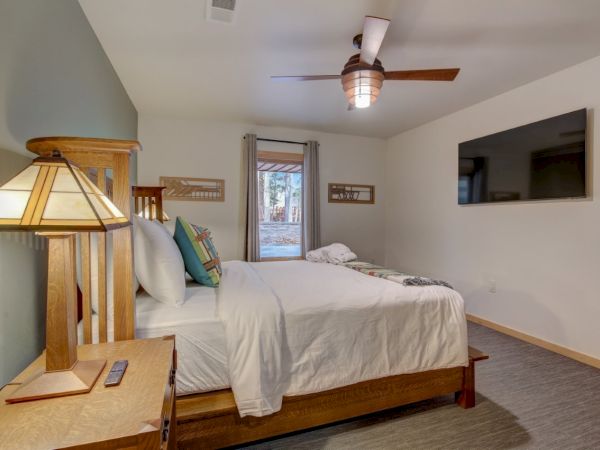 A cozy bedroom with a wooden bed, white linens, colorful pillows, a ceiling fan light, and a window looking out to trees.