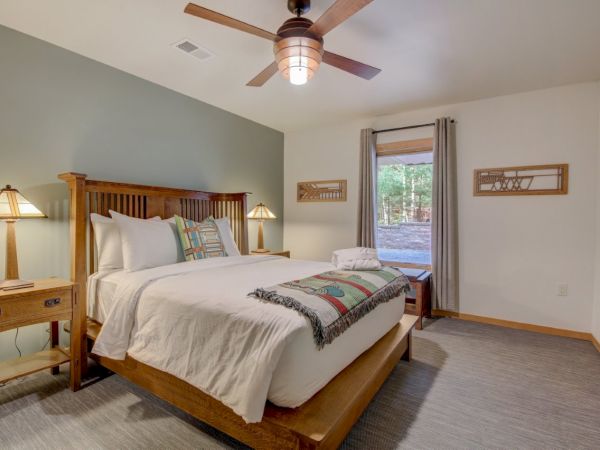 A bright bedroom with a wooden bed, bedside lamps, gray curtains, a window view, and a ceiling fan overhead.