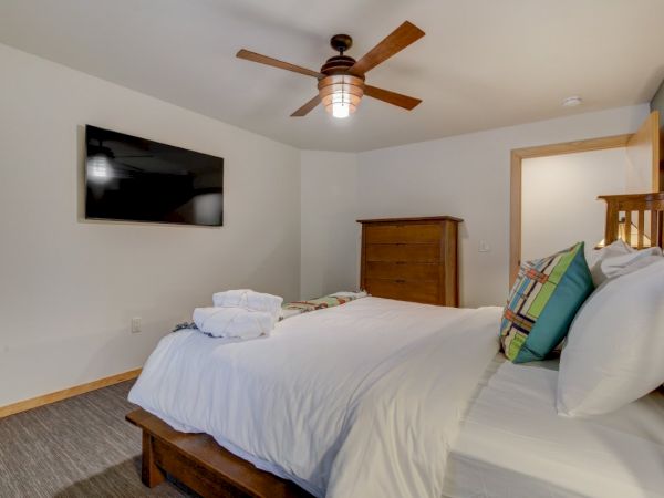 A tidy bedroom with a large bed, wall-mounted TV, wooden dresser, and a ceiling fan overhead.