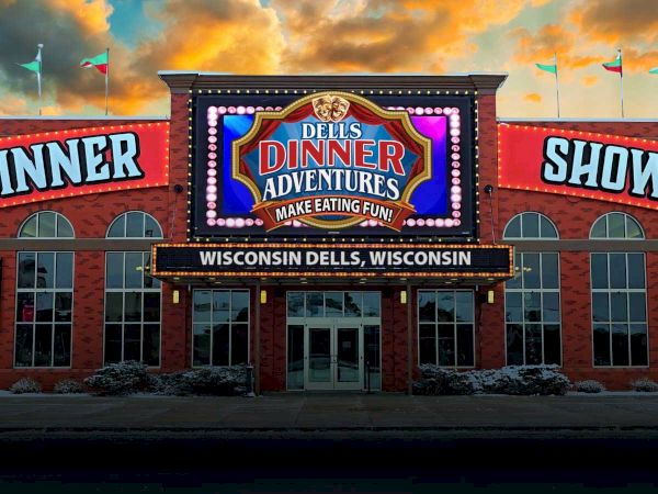 A Wisconsin Dells dinner show marquee reading &ldquo;Dells Dinner Adventures&rdquo; with &ldquo;Make eating fun!&rdquo; lit up at sunset.