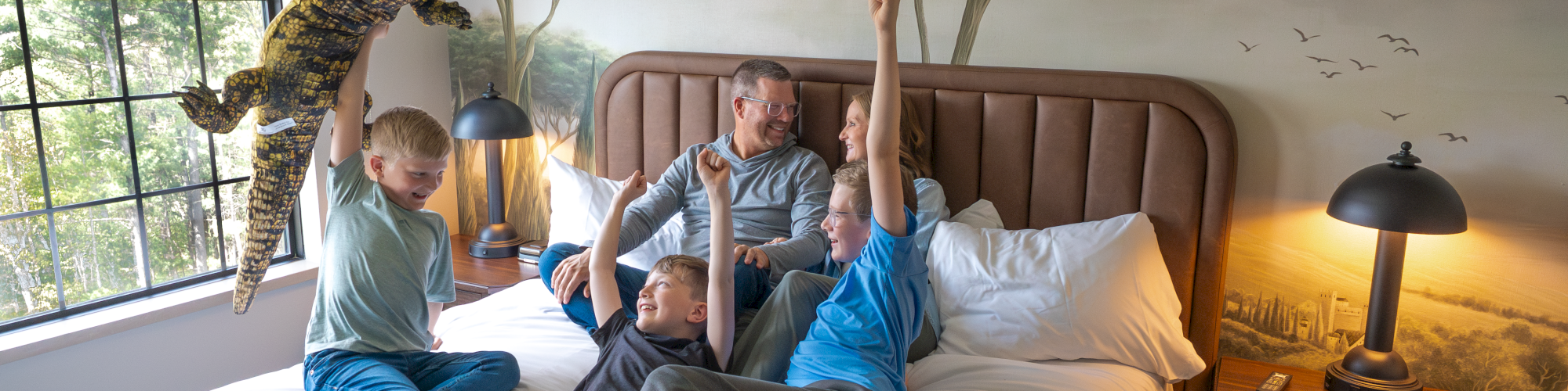 A family plays on a bed in a safari-themed room as a child in a dinosaur suit pretends to roar overhead.